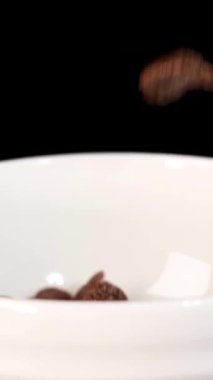 Çikolatalı gevrek taneleri, siyah arka planda beyaz bir kaseye dönüşür. Yavaş çekimde, dinamik ışıklandırmayla yakalanır.
