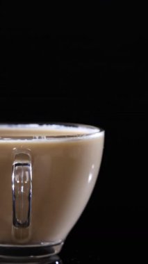 Sütlü kahveyle dolu cam bir bardak siyah bir zemin üzerinde oturur, kremsi dokusunu ve zarif tasarımını vurgular.