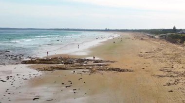İnsansız hava aracı görüntüleri, Avustralya, Ocean Grove 'da yumuşak gün ışığı altında kumlu bir sahil boyunca yürüyen insanları yakalıyor.