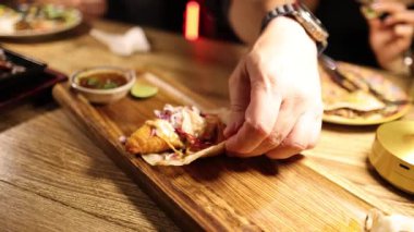 Sıcak ışıklandırma altında ahşap bir tahtanın üzerinde sıcak bir restoran ortamında taco hazırlayan biri.