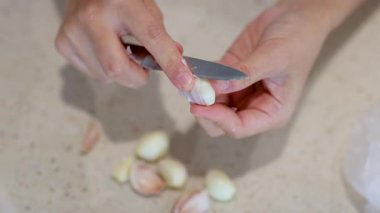 Açık renkli bir mutfak yüzeyinde sarımsak karanfilleri soyan ellerin yakın çekimi. Parlak ışık altında küçük bir bıçak kullanıyorlar.