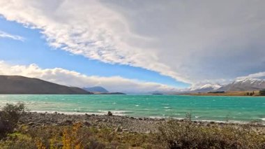 Dalgalı dalgalı turkuaz göl, ön planda dikenli çalılar, karla kaplı dağlar, dramatik bulutlar, ve parlak gün ışığında güçlü rüzgar. Statik geniş açı