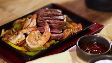 Biftek ve karides fajitalarının yakın plan çekimi, ahşap bir masada soslar, peçetelere odaklanmak için geçiş.