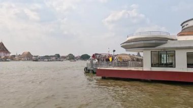 Bir tekne Bangkok 'ta modern bir nehir kıyısındaki binadan geçiyor. Sahne, parlak gün ışığında hareketli nehir yaşamını yakalıyor.