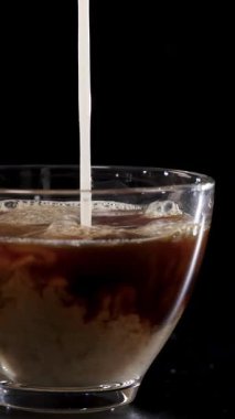 Süt, bir bardak kahvenin içine dökülüyor ve koyu bir arka fonda dönen desenler oluşturuyor.