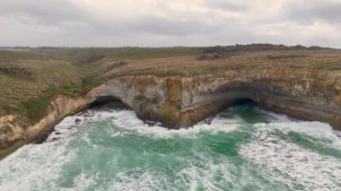 Drone görüntüleri Avustralya, Port Campbell 'ın dramatik uçurumlarını ve turkuaz dalgalarını çekiyor, bulutlu gökyüzü altında, doğal erozyonu ve kıyı güzelliğini vurguluyor.