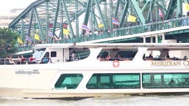 Bir yolcu gemisi Bangkok 'un Memorial Köprüsü' nün altından geçiyor. Canlı bayraklar ve nehir manzarasının keyfini çıkaran yolcular sergiliyor.