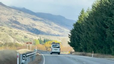 Bir araç Wanaka, Yeni Zelanda 'da, açık gün ışığı altında dağlar ve ağaçlarla çevrili dolambaçlı bir yolda ilerliyor.