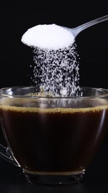 Bir kaşık dolusu şeker, bir bardak çaya dökülür. Koyu bir fonda, granüllerin altını çizer.
