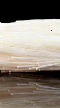 Bir parça enoki mantarı parlak stüdyo ışıkları altında parlak siyah yansıtıcı yüzeyde yavaşça döner, ince detaylar ve pürüzsüz dokular açığa çıkar.