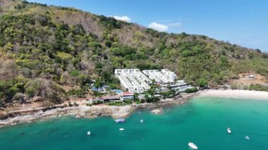 İnsansız hava aracı görüntüleri Phuket, Tayland 'ın sakin sahil şeridini, yemyeşil suları, turkuaz suları ve açık gökyüzü altında demirlemiş tekneleri gösteriyor.