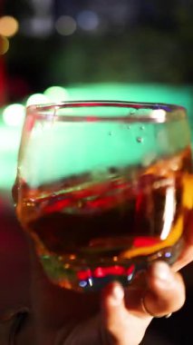 Bir el, bir bardak viskiyi buzla çevirir. Renkli ve odak dışı bar ışıklarıyla aydınlatılır. Dinamik sıvı hareket ve sığ alan derinliği ile yakın çekim.