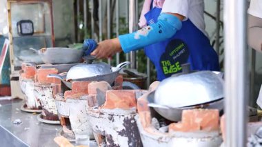 Tedarikçi kalabalık bir Phuket pazarında kilden soba kullanarak geleneksel Tayland atıştırmalıkları hazırlıyor. Parlak ışıklandırma hareketli sokak yemekleri sahnesini vurgular