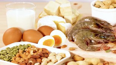 Fındık, süt ürünleri, yumurta ve deniz ürünleri de dahil olmak üzere çeşitli protein zengini yiyecekler parlak ışıklandırmalı ahşap bir yüzey üzerinde sergilenmektedir.