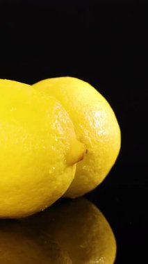 İki koca limon parlak stüdyo ışıkları altında yansıtıcı siyah bir yüzey üzerinde pürüzsüz bir şekilde döner. Parlak sarı renk ve detaylı narenciye dokusunu vurgular.