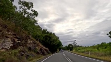 Port Douglas, Queensland, Avustralya 'da, bulutlu gökyüzünün altında, ağaçlarla kaplı, yemyeşil bir otoyol boyunca sakin bir yolculuk.