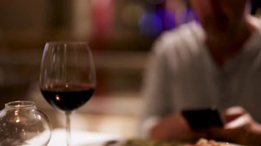 Bir adam lokanta masasında telefonuna odaklanıyor, ön planda bir bardak şarapla.