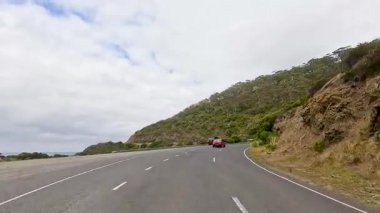 Bir araba dolambaçlı Büyük Okyanus Yolu boyunca yol alır, yemyeşil ve bulutlu bir gökyüzü altında kıyı manzaralarıyla çevrilidir.