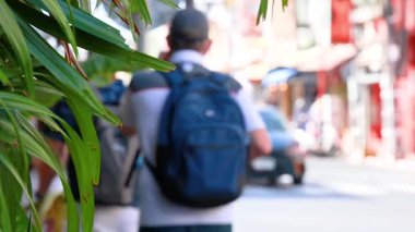 A tourist walks along a bustling street in Phuket, Thailand, surrounded by vibrant colors and lively traffic under bright daylight