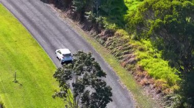 Drone captures a car driving through a lush, green rural landscape under bright sunlight