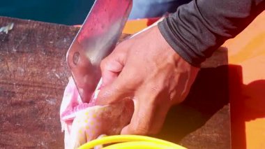 A person skillfully cleans a fish on a wooden board under bright sunlight, using a knife for precise cuts