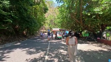 Tourists stroll along a shaded road on Monkey Hill, Phuket, surrounded by lush greenery and bright sunlight