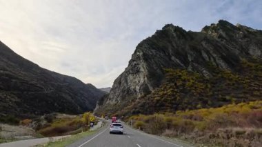 Cars navigate winding roads amidst Queenstown's rugged mountains. Clear skies and natural beauty enhance the serene drive