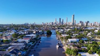 İnsansız hava aracı görüntüleri, açık mavi gökyüzü altında canlı mimari ve yemyeşil alanları vurgulayarak, Gold Coast kentsel ufuk çizgisini ve sakin nehri yakalıyor.