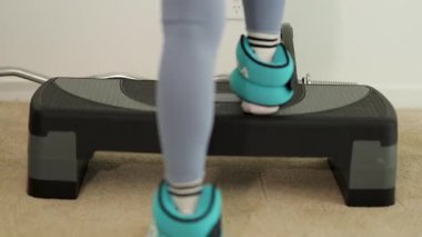 Adım aerobiği yapan kişi, ayak bileği ağırlığıyla bir platformda, kapalı alanda, hafif ışıklandırma altında