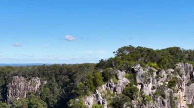 İnsansız hava aracı Nimbin Taşları 'nın panoramik görüntüsünü yakalar, yemyeşil ve parlak mavi gökyüzü altında eşsiz kaya oluşumlarını gösterir.