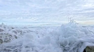 Dinamik okyanus dalgaları kayalık kıyılara çarparak bulutlu bir gökyüzünün altında köpüklü sıçramalar yaratıyor.