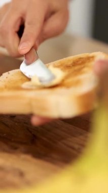 Bir el, sıcak bir mutfakta, bıçakla ekmeğin üzerine tereyağı sürer.