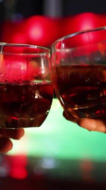 İki el, koyu bir içecekle dolu bardakları rengarenk bar ışıklarına karşı tokuşturur. Sığ alan derinliği, şenlik havası.