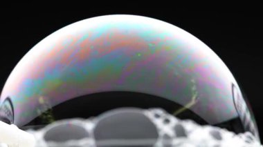 Canlı bir sabun köpüğü, değişen renkleri ve yansımaları karanlık bir arkaplan karşısında gösterir, dinamik ışık oyunu gösterir
