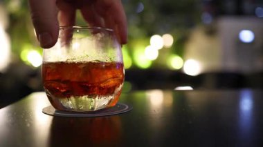 Bir el, koyu bir bar masasındaki bardakta kehribar kokteylini nazikçe kıvırır. Sıcak ortam aydınlatması ve Bangkok 'ta bir gece hayatı ortamında yumuşak bir arka planı vardır.