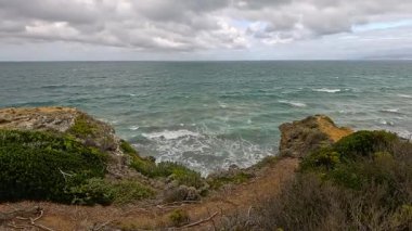 Dalgalar bulutlu gökyüzünün altında engebeli kayalıklara ve kıyı bitkilerine doğru yükseliyor. Statik geniş çekim dinamik okyanus hareketini ve Büyük Okyanus Yolu boyunca dramatik doğal manzarayı yakalar.