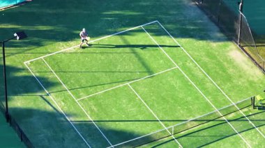 Drone, güneşli bir kortta topa vuran bir tenisçiyi yakalıyor, dinamik hareket ve canlı renkler sergiliyor.