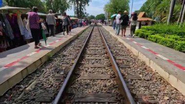 Yayalar, parlayan bir açık hava pazarındaki demiryolu raylarının yanında, yeşil ve satıcı tezgahları platformu kaplarken, gün ışığında ilerliyorlar. Statik, göz hizasında çekim