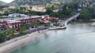 Drone görüntüleri Phuket, Tayland 'da kıyı şeridi boyunca verimli yeşillik ve canlı mimari gösteren manzaralı bir sahil beldesi yakalıyor.