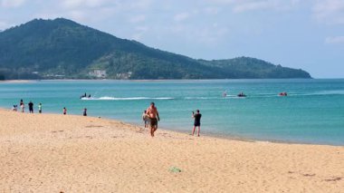 Turistler açık gökyüzü ve arka planda uzak jet ski 'lerle güneşli bir Phuket plajı boyunca yürürler.