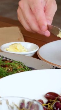 Bir insan kahvaltı masasında bir dilim rustik ekmeğe tereyağı sürmek için tereyağı bıçağı kullanır, doğal ışıklandırma ve yakın çekim kamera açılarıyla.