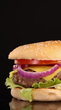 Peynirli, marullu, domatesli ve soğanlı sulu bir sığır burger parlak stüdyo ışıkları altında yansıtıcı siyah bir yüzey üzerinde pürüzsüz bir şekilde döner.