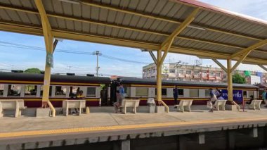 Bir yolcu treni Kanchanaburi istasyonundan peronda ve vagonların içinde gezginlerle birlikte kapalı bir platform altında yavaşça ayrılır. Gün ışığı, sabit geniş açı