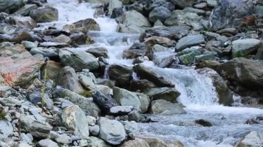 Açık bir dağ deresi engebeli bir doğa ortamında geniş kayaların üzerinden hızla akar. Doğal gün ışığı manzarayı aydınlatır. Kamera sabit duruyor.