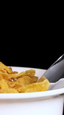 Bir kaşık beyaz bir kaseden mısır gevreğini kaldırır, siyah bir zemine yaslanır, mısır gevreğinin dokusunu ön plana çıkarır.