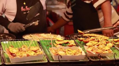 Tedarikçiler hareketli bir pazarda şiş ızgara yapıyor, hareketli sokak yemekleri kültürünü canlı etkileşimler ve sıcak ışıklandırmayla vurguluyorlar.