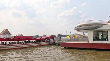 Bangkok 'ta bir nehir manzarası bulutlu bir gökyüzünün altında hafif su hareketleriyle bir rıhtımı, binaları ve tekneleri gösteriyor.