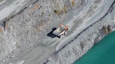 Telefoto İHA, aydınlık gün ışığında turkuaz bir göl kenarındaki taş ocağında yol alan kamyonları yakalıyor.