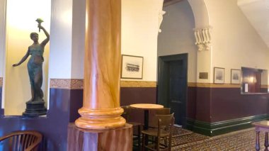 Klasik dekorlu bir otel lobisi, içinde bir heykel, ahşap sütunlar ve sıcak ışıklandırma olan davetkar bir atmosfer yaratıyor.