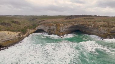 Drone görüntüleri Avustralya 'daki Port Campbell' da dramatik kayalıkları ve çarpışan dalgaları yakalar. Bulutlu gökyüzü engebeli, doğal güzelliği arttırır.